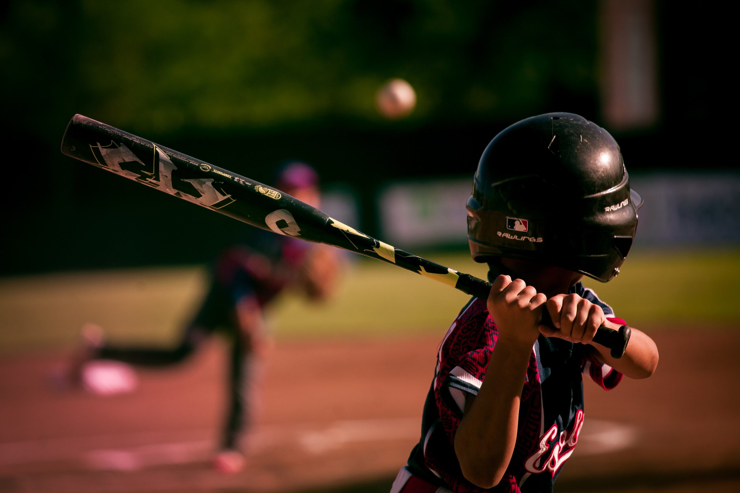A young baseball player wearing a helmet prepares to swing at a pitch, while another player in the background throws the ball on a sunny field.