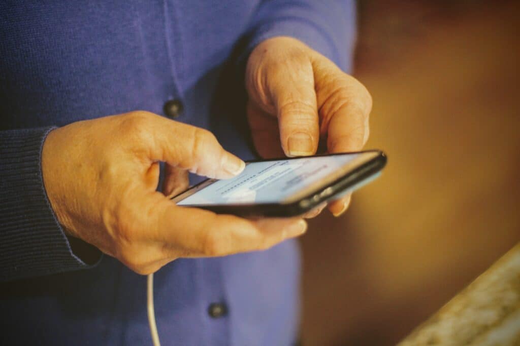 A person wearing a blue sweater uses a smartphone. The image captures their hands holding the device, focused on the screen, while an off-white background is slightly visible.