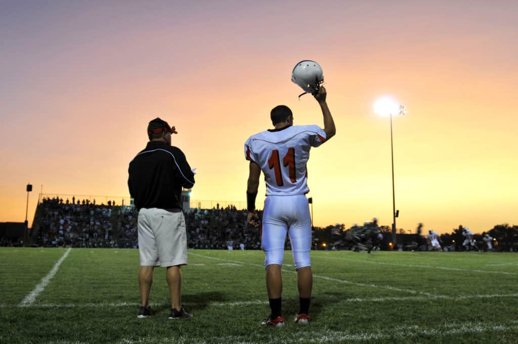 Football player in a white uniform holds his helmet up, standing next to a coach on the sidelines. The sun sets in the background, casting an orange glow over a crowded stadium during a game.