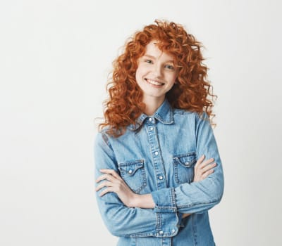 Person with curly red hair smiling, wearing a denim shirt, arms crossed, against a plain background.