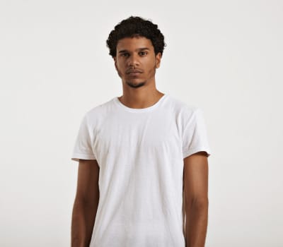 A person with short curly hair is wearing a plain white t-shirt and standing against a neutral background.