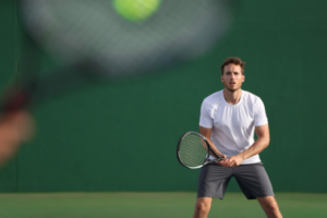 A man in a white shirt and gray shorts stands ready with a tennis racket on an outdoor wiregrass tennis court, preparing to receive a serve. The blurred racket of his opponent is visible in the foreground.